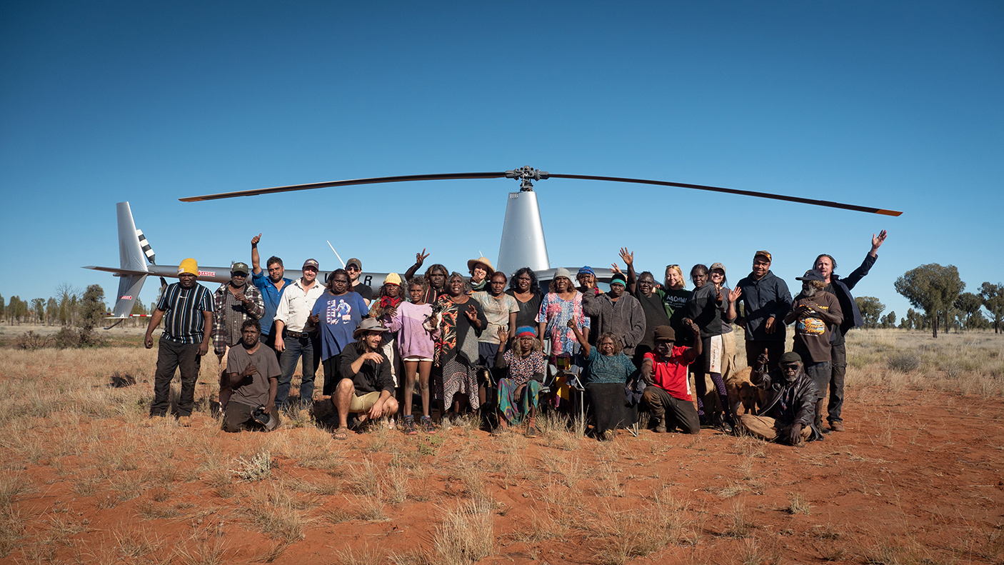 A large group of people at the Tjunukutjarra camp stand in a desert environment waving at the camera