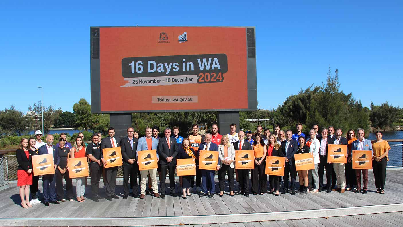 A large group of athletes stand with government and sports industry leaders to stand up to violence against women. The Swan River and a large 16 Days in WA billboard is in the background.