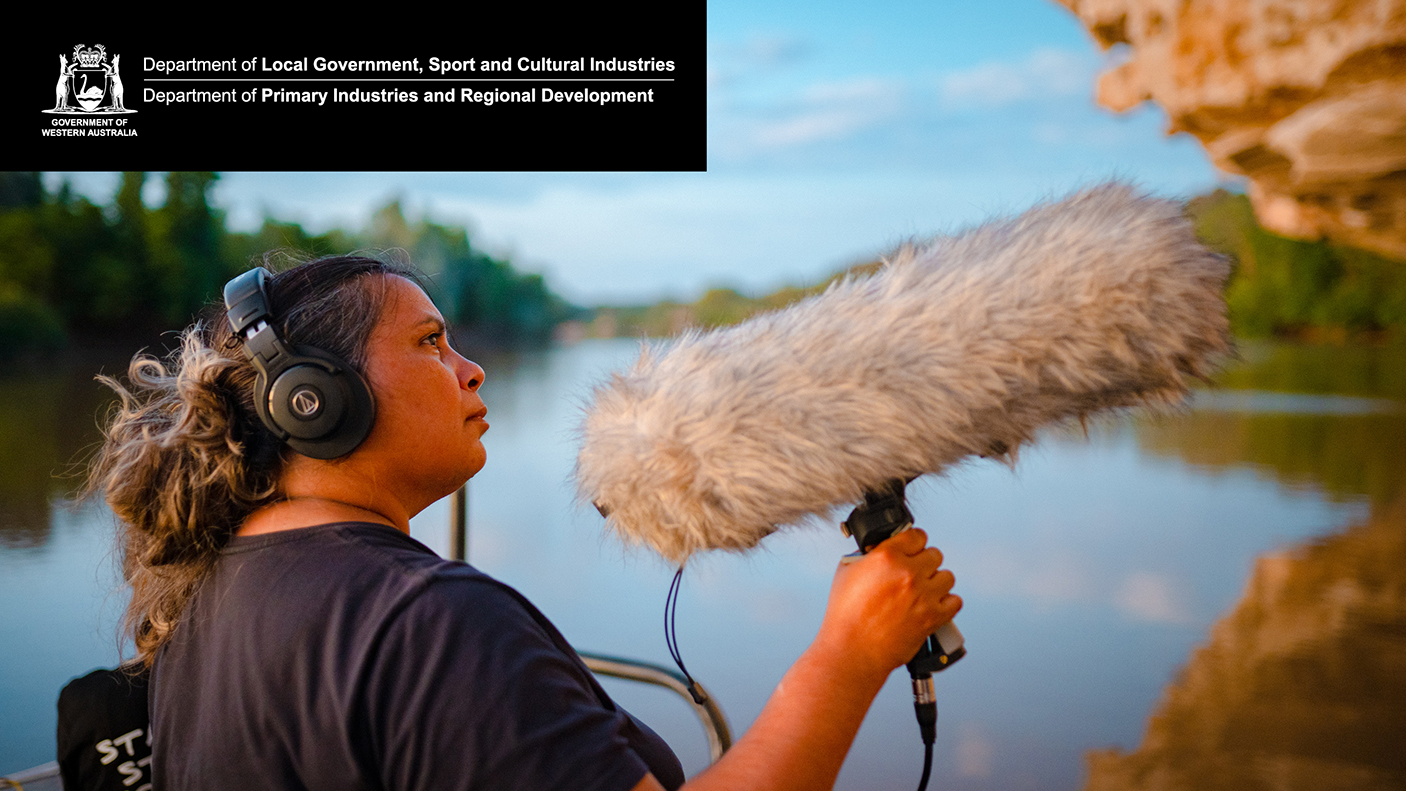 Sound technician wearing headphones holds up a microphone covered with a windsock. A regional river is in the background.