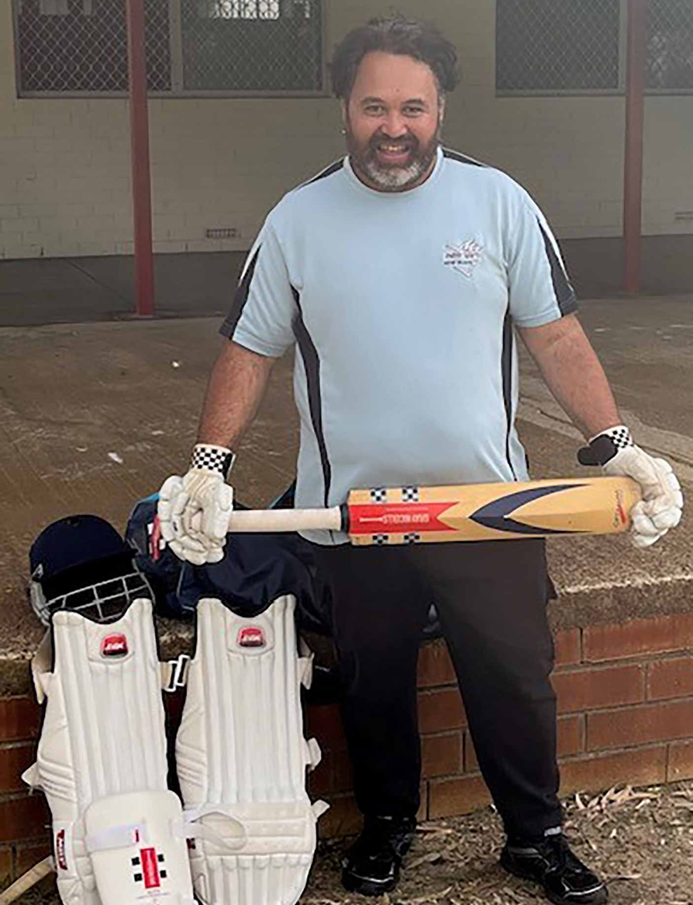 A man stands holding a cricket bat, wearing crick gloves, next to his helmet and shin guards