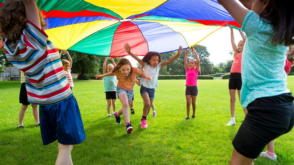 Young children playing in a grassy park, and running under a large colourful sail shade